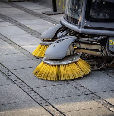 Close-up view of a road sweeper's yellow brushes on a paved street.