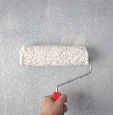 A person's hand using a paint roller to apply white paint on a wall.